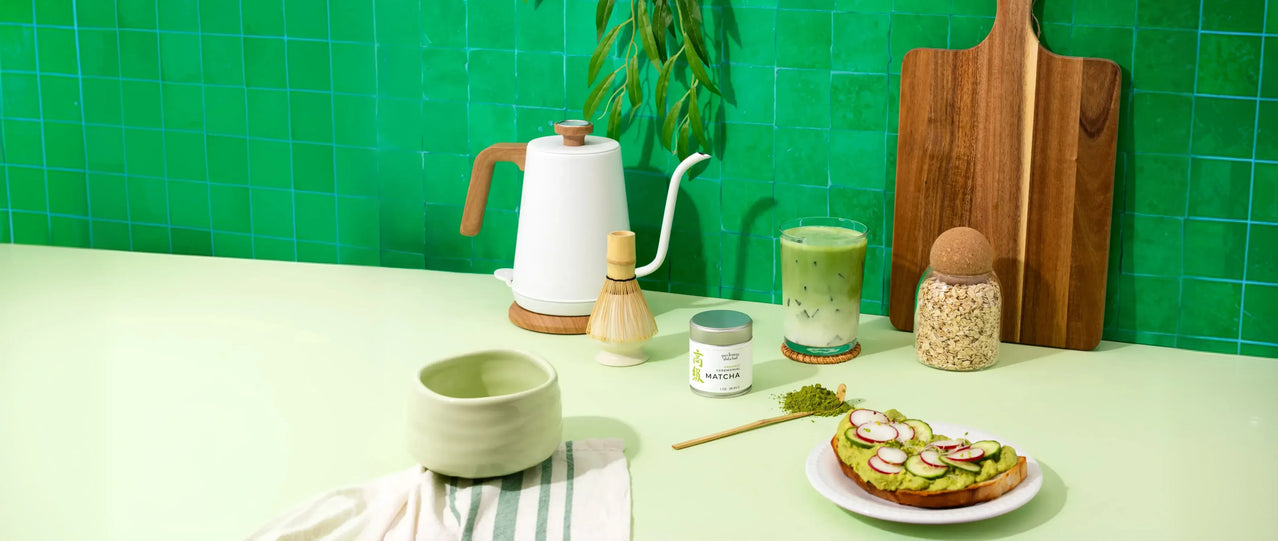 Tea-making setup with a kettle, cups, and a plate of food on a green tiled wall background.
