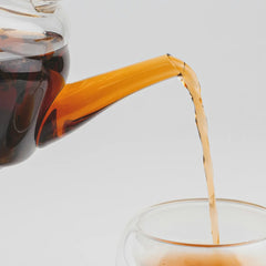 Glass teapot with a yellow handle pouring tea into a glass cup on a light gray background