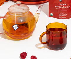 Tea set with a teapot, mug, and box of Organic African Sunset tea on a white surface.