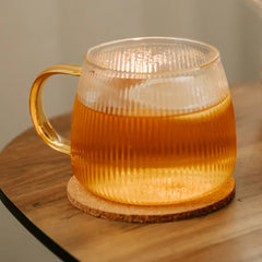 Glass mug with a textured handle filled with a warm beverage on a wooden coaster.