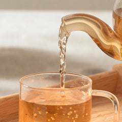 Tea being poured from a glass teapot into a clear glass mug on a wooden surface.