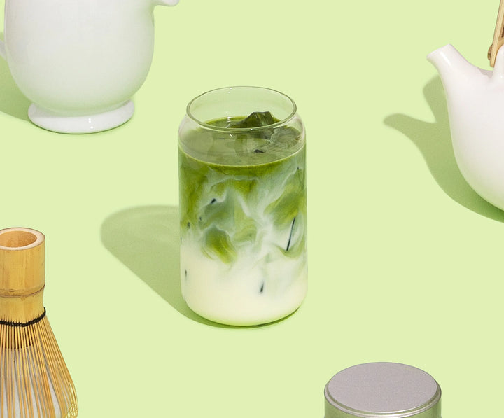 Glass of iced matcha tea with a white teapot and bamboo whisk on a light green background